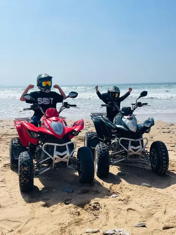 Helmeted riders with red and black quad bikes beside the sea